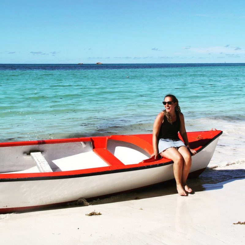 Sentada em um barco, Amanda posa com a praia de água cristalina ao fundo, em Seychelles.