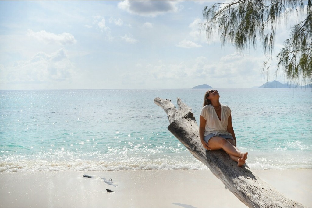 Em frente à praia de água cristalina, em Seychelles, Amanda usa um tronco seco e grande de uma árvore como banco para admirar a paisagem.