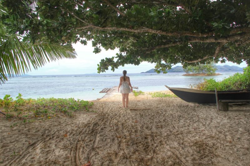 De frente para o mar, Amanda Noventa aparece de costas, ao lado de uma pequena embarcação, coberta por árvores.