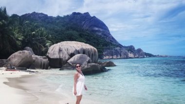 Amanda posa sorridente em frente às grandiosas pedras que ficam na orla da praia de água cristalina Anse d´Argent, em Seychelles.
