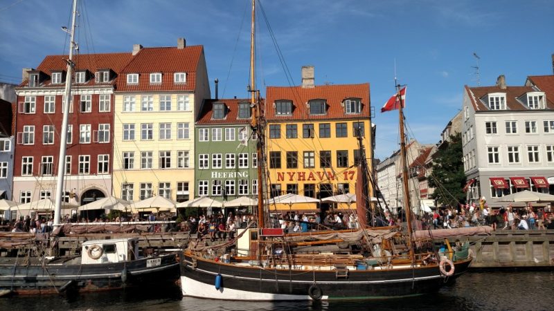 Foto do de frente para o pier, com embarcações atracadas diante das mesas ao ar livre dos bares e restaurantes de Nyhavn, em Copenhagen, Dinamarca.