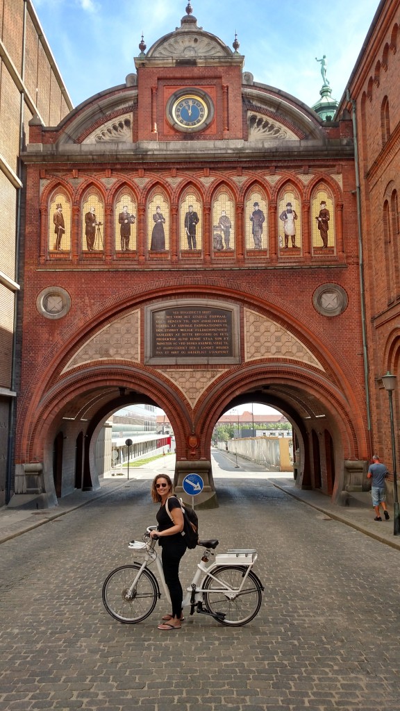 Segurando a bicicleta, Amanda sorri em frente a fachada da Cervejaria Carlsberg, em Copenhagen, Dinamarca.