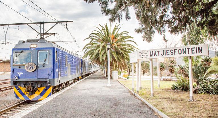 Estação de trem na África do Sul, com trem azul chegando.