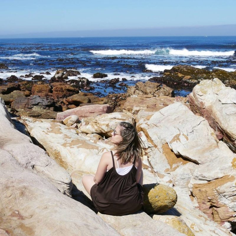 No Cabo da Boa Esperança, Amanda desfruta da paisagem posando para foto em pedras acessíveis durante a maré baixa.