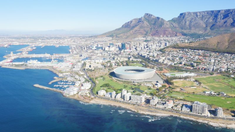 Cape Town vista de um helicóptero, com paisagem que engloba mar banhando a orla de prédios, estádio de futebol da cidade e Table Mountain ao fundo.