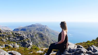 No topo da montanha Table Moutain, Amanda posa para foto sentada em uma pedra enquanto admira a paisagem formada pelo oceano se confundindo com as nuvens ao fundo.