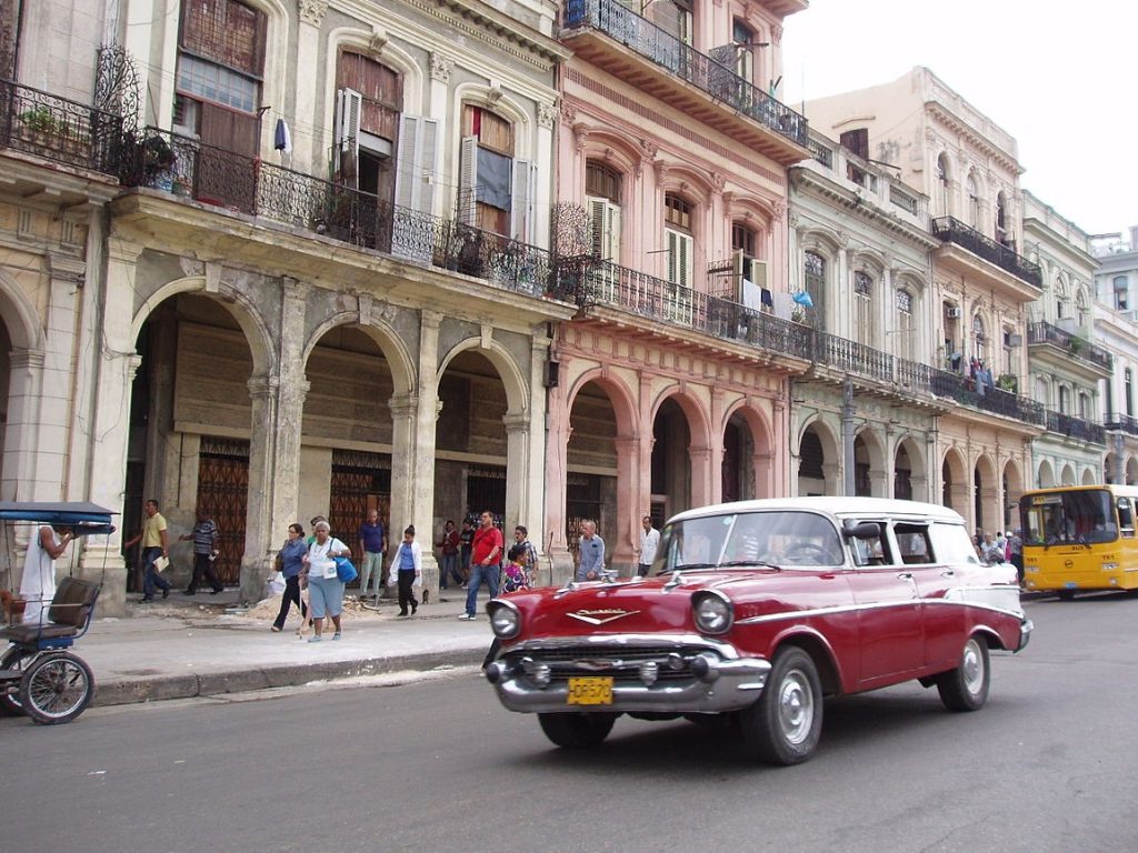 Rua em Habana Vieja com um carro antigo em destaque, pedestres caminhando pela calçada, e uma fachada de prédios antigos ao fundo.