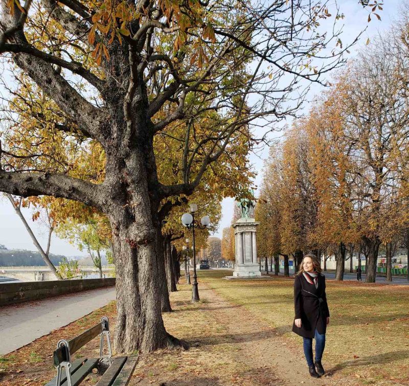 Amanda posa para foto em praça de Paris, às margens do rio Sena. As folhas laranjadas das árvores indicam que é outono.