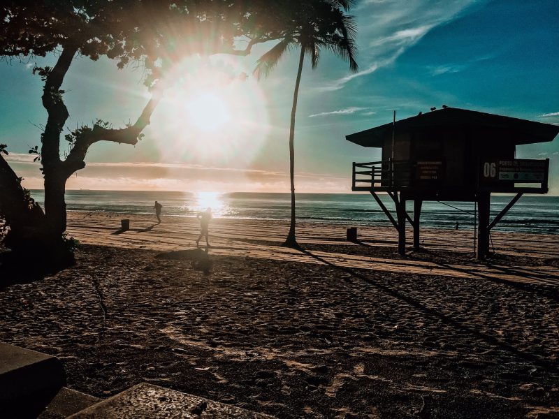 Amanhecer na Praia de Boa Viagem, em Recife, com destaque para orla com pessoas caminhando, uma árvore e um quiosque de salva-vidas às margens.